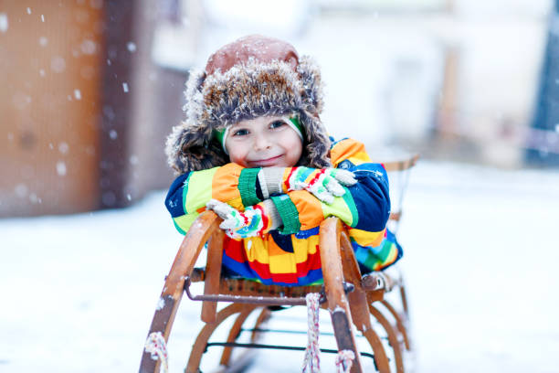 Little kid boy enjoying sleigh ride during snowfall. Happy preschool kid riding on vintage sledge. Child play outdoors with snow. Active fun for family Christmas vacation in winter stock photo