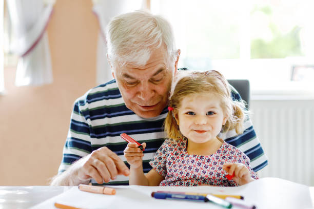 Cute little baby toddler girl and handsome senior grandfather painting with colorful pencils at home. Grandchild and man having fun together stock photo