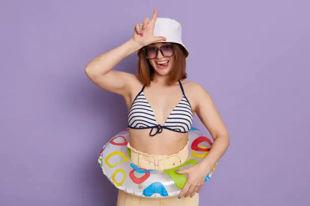 Portrait of Caucasian dark haired woman wearing swimsuit and panama, showing looser gesture, looking at camera, posing with rubber ring isolated over purple background.