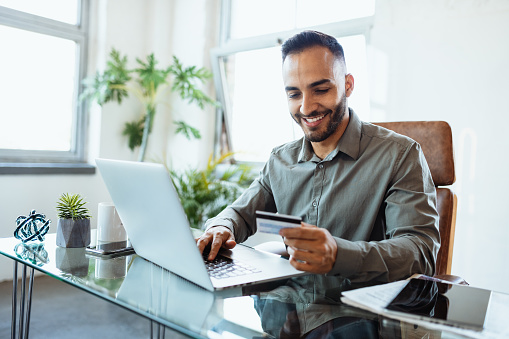 Hombre de negocios latino sonriente en la oficina, usando tarjeta de crédito para pagar en línea photo