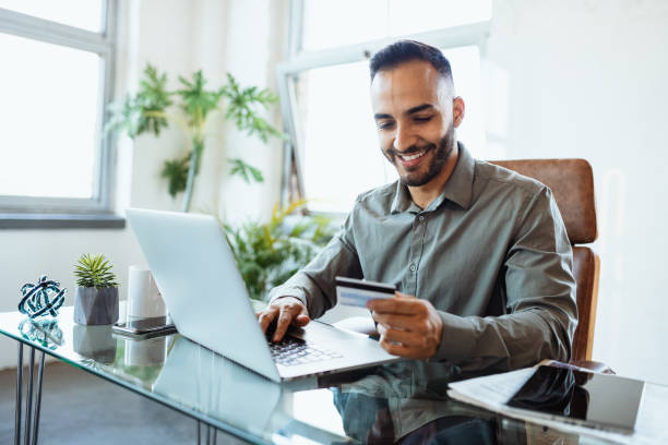 hombre de negocios latino sonriente en la oficina, usando tarjeta de crédito para pagar en línea - pagar fotografías e imágenes de stock