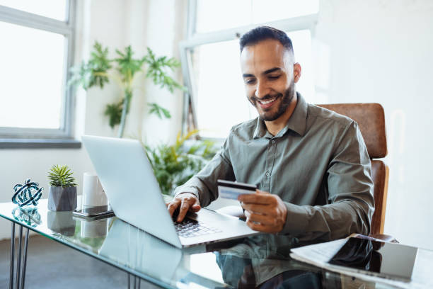 uomo d'affari latino sorridente di metà adulto in ufficio, usando la carta di credito per pagare online - pagare foto e immagini stock