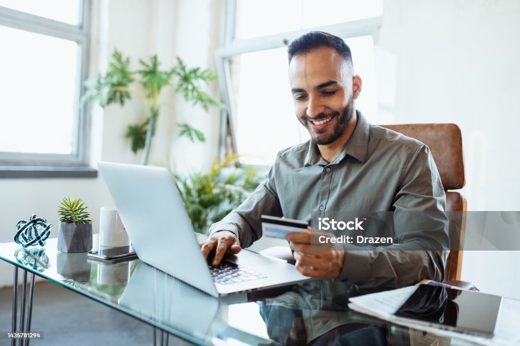 Mid adult smiling Latin businessman in office, using credit card to pay online Mid adult Latin businessman in office, working on laptop Credit Card Stock Photo