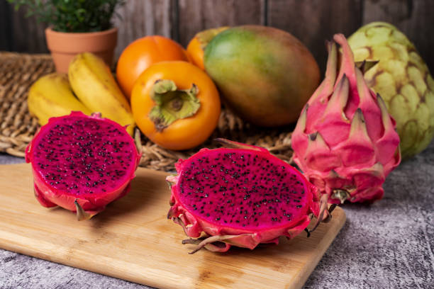 istock Pitahaya or dragon fruit split in two on a wooden board, exposing the pink interior and black seeds, in the background some tropical fruits such as cherimoya, mango, persimmon and bananas, foreground. 1435785604