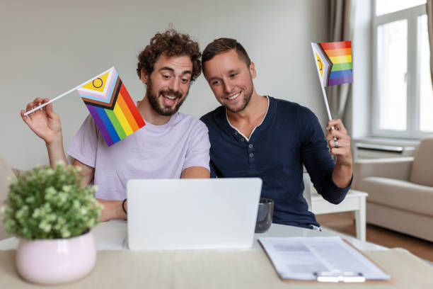 Male lovers video calling their friends in their living room at home. Young gay couple smiling cheerfully while greeting their friends on a video call. Holding LGBTQ flags and celebrating pride month together Male lovers video calling their friends in their living room at home. Young gay couple smiling cheerfully while greeting their friends on a video call. Holding LGBTQ flags and celebrating pride month together LGBT Online Therapy stock pictures, royalty-free photos & images