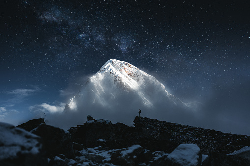 Brave solo mountain traveler stand in front of huge snowy mountain and starry sky. Professional tourist on mountains summit looking on greate snow cloudy rock