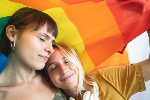 Portrait of young lesbian couple hugging each other while standing together under the rainbow flag