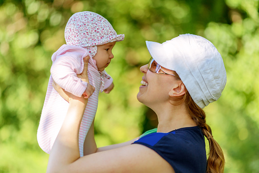 Newborn baby outdoor, happy mother holds her small child in hands. Cute infant in pink on blurred green trees background. Young woman and her baby girl in sunny park or garden. Happiness concept.