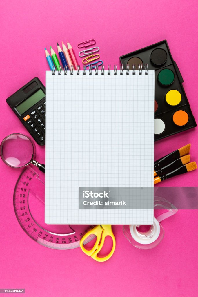 Office and School Supplies Directly above view of a desk full of office and school supplies like watercolor, paper clip, colored pencil, magnifier, calculator, paintbrush, scissors, ruler and a  checkered notebook in the middle on pink background. Adhesive Tape Stock Photo