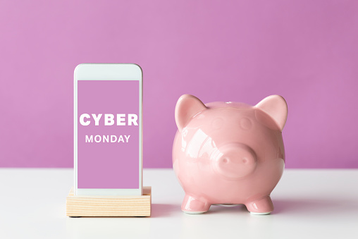 Front view of a smart phone and pink piggy bank on white table in front of a purple background. Cyber Monday text on device screen.