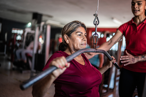 Senior woman pulling weight machine with help of fitness trainer at the gym