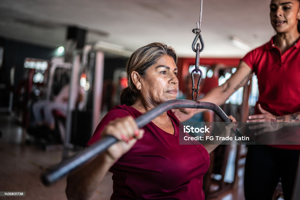Senior woman pulling weight machine with help of fitness trainer at the gym Exercising Stock Photo