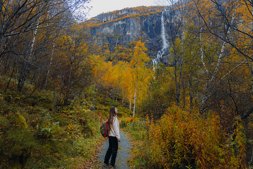 Happy woman with long hair, with backpack and in fleece jacket walking at the beautiful footpath overlooking the yellow woodland and the waterfall in Jostedalbreen National park, Western Norway