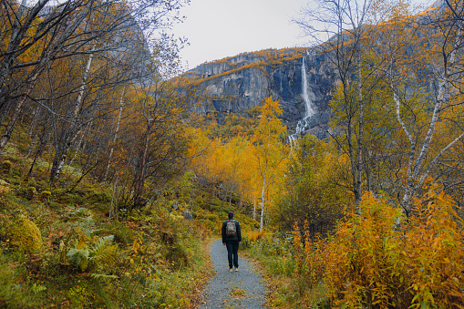 Happy man with backpack and in jacket walking at the beautiful footpath overlooking the yellow woodland and the waterfall in Jostedalbreen National park, Western Norway