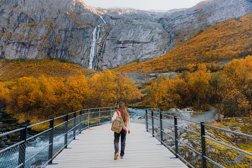 Happy woman with long hair, with backpack and in fleece jacket walking at the beautiful bridge overlooking the yellow woodland and the waterfall in Jostedalbreen National park, Western Norway