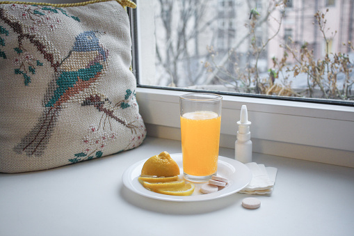 Seasonal cold: glass cup with liquid vitamin C, sliced lemon, instant pills and nasal spray with paper napkins on the windowsill. Next to the vintage embroidered pillow