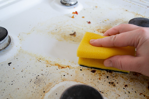 A hand with a yellow wash sponge washes the very dirty greasy surface of the gas stove. After the sponge, a clean trace remains.