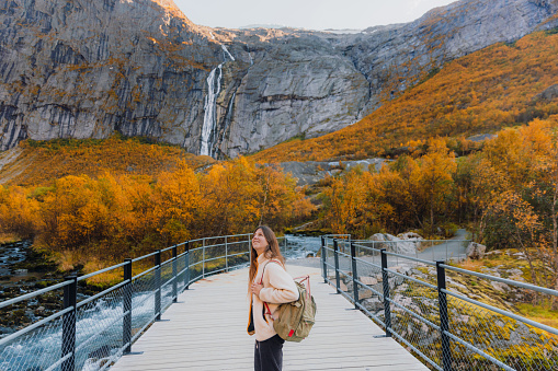 Happy woman with long hair, with backpack and in fleece jacket walking at the beautiful bridge overlooking the yellow woodland and the waterfall in Jostedalbreen National park, Western Norway