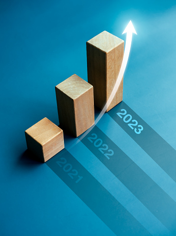 Shining rise up arrow on wooden cube blocks, bar graph chart steps on blue background with year numbers, 2023, 2022, 2021, vertical style. Business growth and development to success concepts.