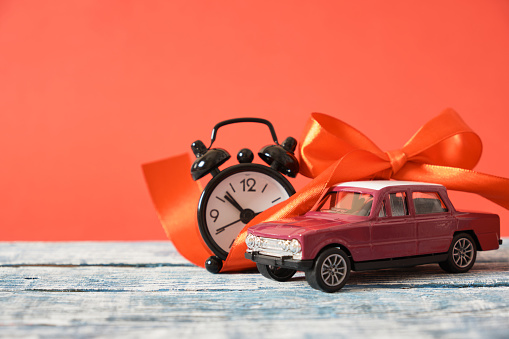 miniature purple retro car model, red ribbon bow and alarm clock on wooden surface, copy space, red background