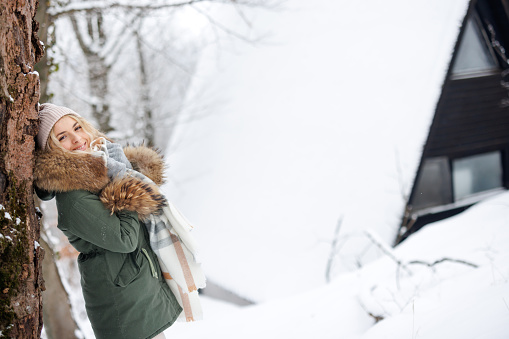 Portrait of young woman leaning on tree, looking at camera and smiling in snowy forest on cold winter day