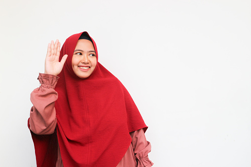 Beautiful asian muslim in hijab looking up and wave hand to greeting, isolated on white background