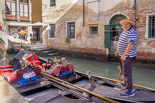 Venice, Italy - October 7th 2022: Mature and traditional dressed gondolier standing in his gondola in a narrow canal in the center of Venice