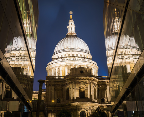 The historic dome of St. Paul’s Cathedral spotlit at night reflecting in the modern glass facade below in the heart of the City of London, UK.