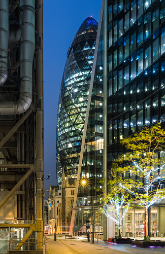 The iconic curves of 30 St Mary Axe illuminated at night overlooking the historic tower of St Andrew Undershaft Church beside the Lloyds Building in the heart of the City of London Financial District, UK.