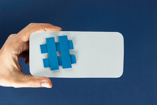 Hand is holding a blue hashtag symbol attached to a white smartphone like shape in front of blue background