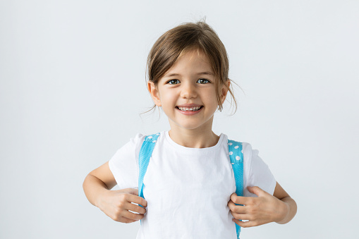 Caucasian little girl with blue backpack with a toothy smile is looking at camera in front of white background.