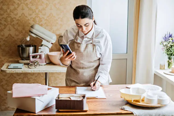 Small local business owner of confectionery, sweet-shop, pastry-shop. Asian Arabic woman pastry chef checking orders in notebook and on phone