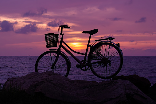 a silhouette of a foot push bicycle in a purple and golden sunset parked near the ocean rocky mountain. Sri Lanka