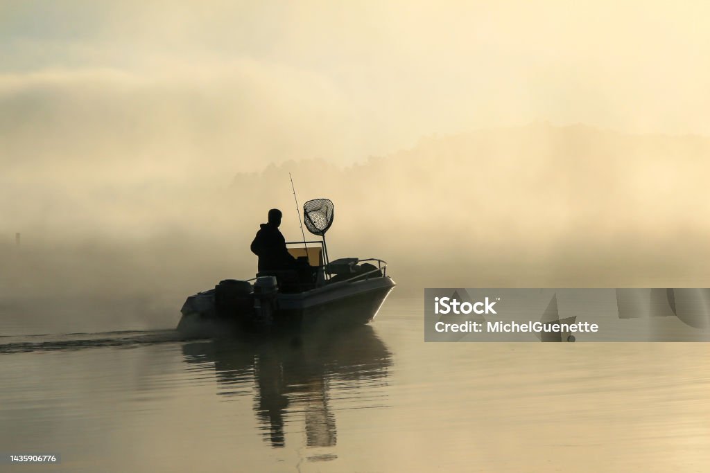Early morning fisherman with fog on the lake. Silhouette of a fisherman in his boat, early in the morning with a lot of fog on the lake. Fishing Stock Photo