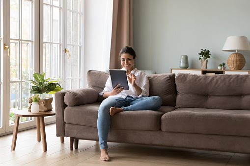 Pretty Indian woman sit on sofa holds digital tablet, enjoy weekend leisure alone at home with electronic device using new application, buying goods on-line, make easy remote order. Modern tech usage