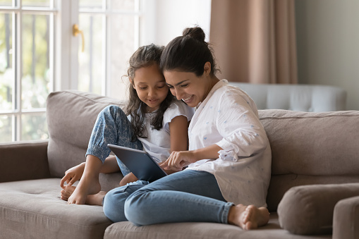 Little cute Indian girl and mom use digital tablet, resting together at home, buying goods through retail services webstore, learn new amusing application seated on cozy sofa, enjoy modern tech usage