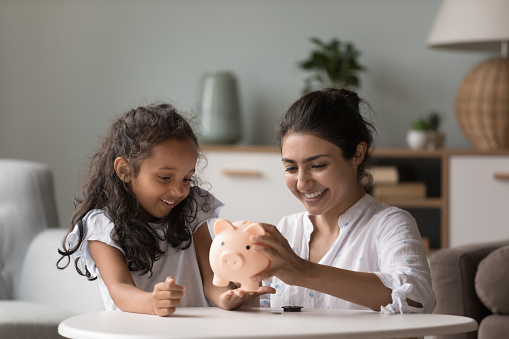 Young happy Indian woman and preschooler daughter sitting at table in living room take out money stack from piggy bank for spending money for future purchases, smile enjoy moment. Savings, finances