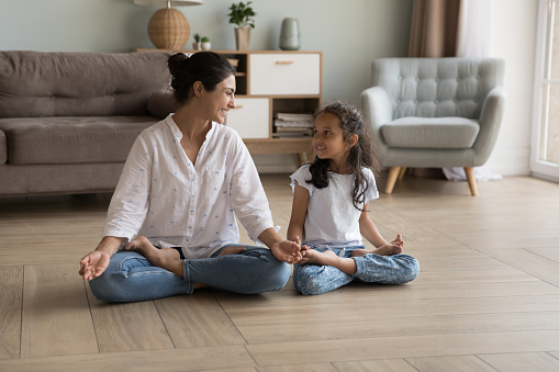 Indian woman laughing seated cross-legged on floor doing meditation practice with cute little daughter at modern cozy home. Loving parent teach child yoga, instill good habits for healthy lifestyle