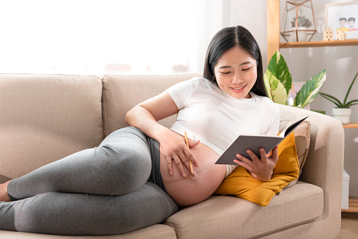 beautiful asian pregnant woman resting on sofa reading a book in afternoon stroking her belly affectionately.