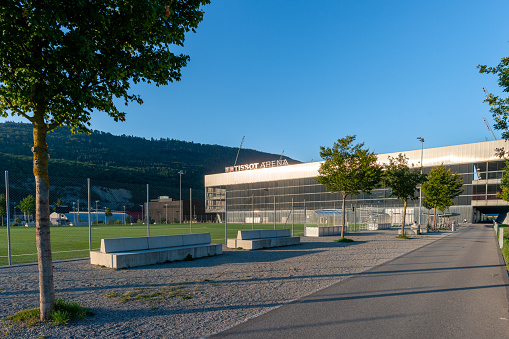 Exterior looking across the green outdoor sports fields to the Tissot Arena building. There is an ice rink in the arena where the ice hockey club EHC Biel plays. and a football stadium where the football club FC Biel-Bienne 1896 plays. The building also houses the Shopping Les Stades shopping centre. 08/28/2022 - Boulevard des Sports 18, 2504 Biel Bienne, canton Bern, Switzerland