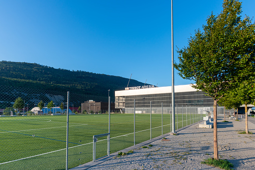 Exterior looking across the green outdoor sports fields to the Tissot Arena building. There is an ice rink in the arena where the ice hockey club EHC Biel plays. and a football stadium where the football club FC Biel-Bienne 1896 plays. The building also houses the Shopping Les Stades shopping centre. \n08/28/2022 - Boulevard des Sports 18, 2504 Biel Bienne, canton Bern, Switzerland