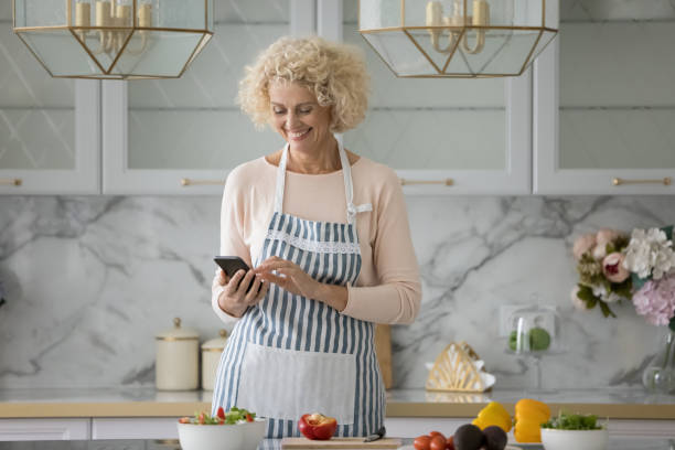 Older woman use smartphone standing alone in modern kitchen