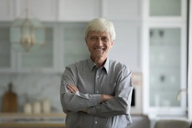 Retired grey-haired man standing in fashionable domestic kitchen with arms-crossed smile look pose for camera. Happy homeowner, wellbeing carefree life on retirement, optimistic older person portrait