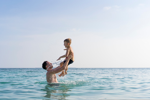 Dad and son are playing in the sea. The father tosses the son up. Maldives islands, water splashes, vacations, blue sky and sunny day.