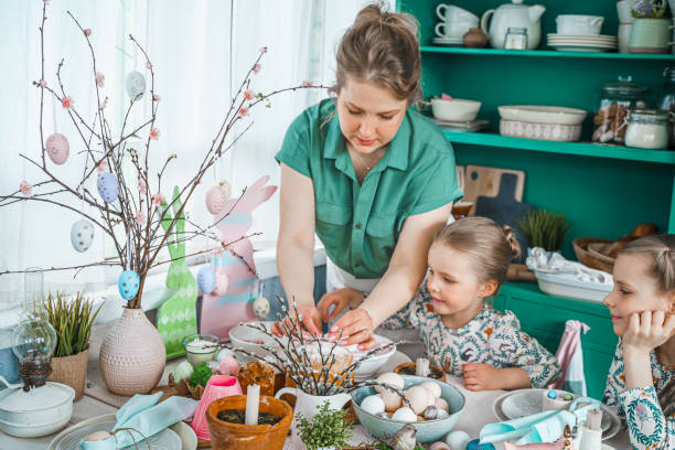 istock Girls, sisters, mother at table with Easter decoration. Celebration in kitchen.Tablescape for home Easter holiday.Family religious traditional festive food, meal. Colored eggs,cake, fun, bunny ears 1435949601