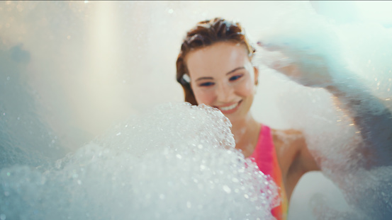 Beautiful woman in swimsuit resting in a bubble bath. Covered in foam and playing with bubbles. Getting pampered in the spa and practicing wellness.
