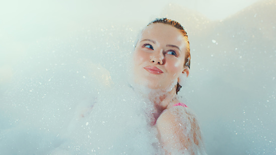 Beautiful woman resting in a bubble bath. Covered in foam and flirting with the camera. Getting pampered in the spa and practicing wellness.