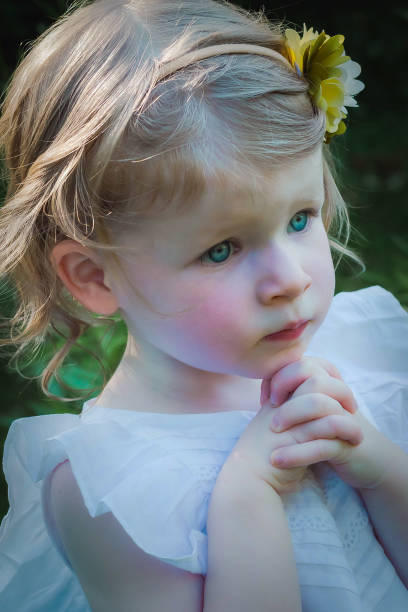 Toddler girl with a emotional expression and folded hands, wearing a white dress and flower headband