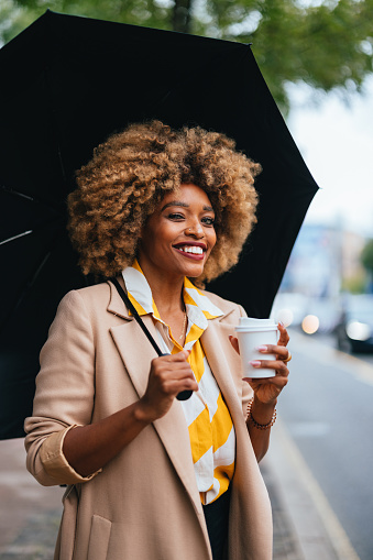 Portrait of a beautiful businesswoman standing on the city street holding a cup of coffee to go and an umbrella. She is smiling and looking away like she is recognizing someone.