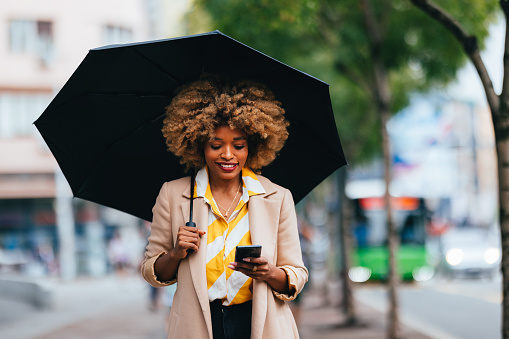 Beautiful and elegant African American businesswoman, walking down the city street with an umbrella, smiling and reading a message on her mobile phone.